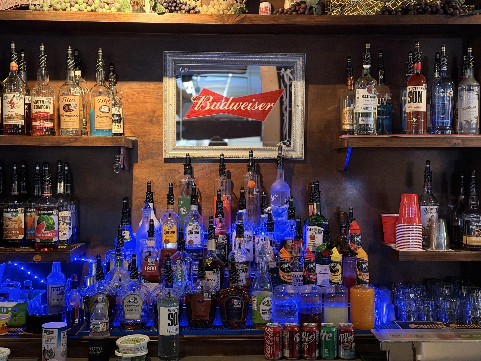 The back bar lined with liquor bottles under blue light and a Budweiser mirror